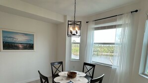 Een eetgedeelte met een ronde tafel, vier stoelen, een kroonluchter, een schilderij aan de muur en een raam met gordijnen.