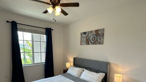 Een slaapkamer met een bed, een plafondventilator, een raam met gordijnen, een nachtkastje met een lamp en een kunstwerk aan de muur.
