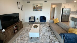 Een woonkamer met een flatscreen televisie, een salontafel, twee fauteuils, een bank en een keuken met een koelkast.