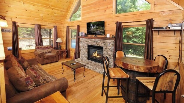 Interior acogedor de una cabaña con chimenea de piedra, mobiliario de madera y ventanas amplias que ofrecen vistas al exterior.