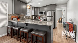 A modern kitchen with dark gray cabinets, a granite countertop, and stainless steel appliances. There are three wooden bar stools and a small island.