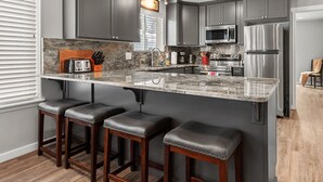 A modern kitchen with dark gray cabinets, a granite countertop, and stainless steel appliances. There are three wooden bar stools and a small island. - Newly Remodeled Home Across from Golf Course—On Centennial Trail, Near Elke Point Wedding Venue (Coeur d'Alene)