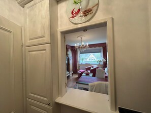 A room with a large mirror reflecting a dining area, a chandelier, and a window with a view of a house.