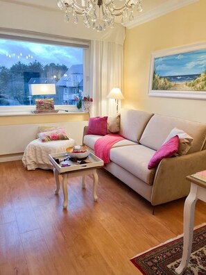 A cozy living room with a beige sofa, a small white table, and a window with a view of a building. - Sunapp - Individuell Ausgestattetes Appartement (Sylt)