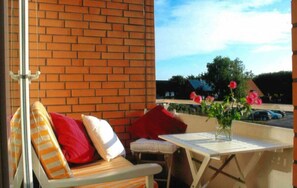 A balcony with a brick wall, a wooden bench with red cushions, a small table, and a vase with flowers.