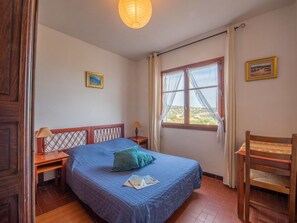 Un dormitorio con cama, ventana con cortinas, una silla de madera, mesita de noche y lámpara.