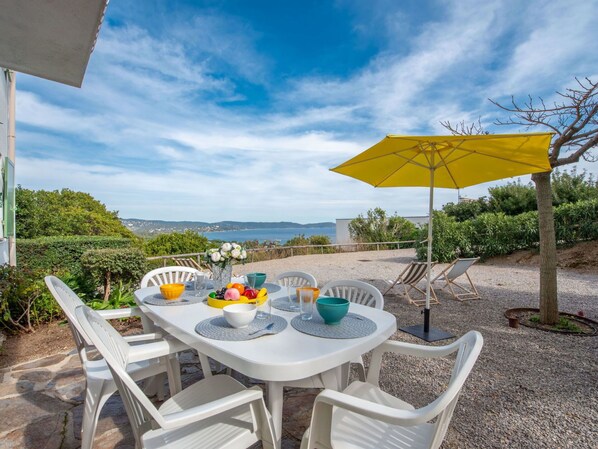 Ein Essbereich im Freien mit einem Tisch für vier Personen, umgeben von weißen Stühlen, einem gelben Sonnenschirm und Blick aufs Meer.