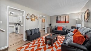 Ein modernes Wohnzimmer mit einer schwarzen Ledersofa, einem Couchtisch und einem geometrischen Teppich. Im Hintergrund ist eine Küche mit weißen Schränken und einer Mikrowelle erkennbar.