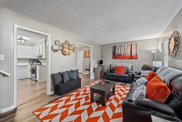 A modern living room with a black leather sofa, a coffee table, and a geometric rug. There is a kitchen visible in the background with white cabinets and a microwave. - Cozy Home with everything you need at your finger tips. (Augusta)