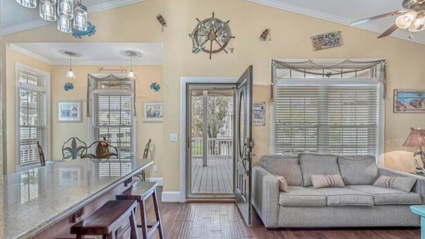 A spacious living area with a large window, a sofa, and a wooden bar counter. - Tiki Hut W/Tiki Bar|Linens|Golf Cart|Outdoor Area (Myrtle Beach)
