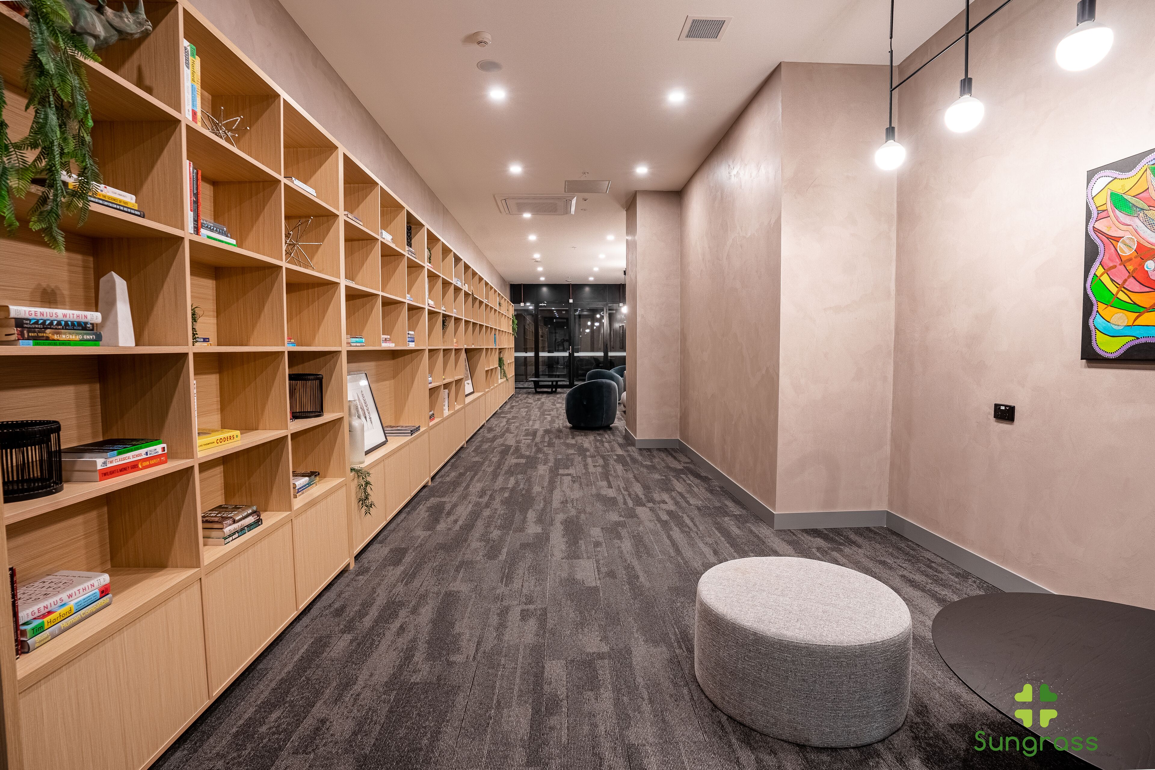 A modern hotel lobby with a carpeted floor, wooden bookshelves, a round seating area, and a colorful artwork on the wall.