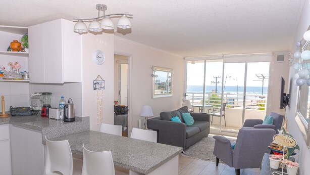 Une cuisine moderne avec des meubles blancs, un plan de travail en granit et un coin repas avec des chaises blanches. Le salon est doté d’un canapé gris avec des coussins bleus, et un balcon offre une vue sur la mer.