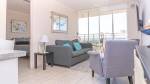 Un salon avec un canapé gris, une petite table avec une lampe, un fauteuil gris et une grande fenêtre donnant sur la mer.
