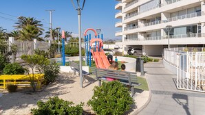 Un terrain de jeux avec un toboggan et des bancs, entouré d’une clôture et de palmiers.