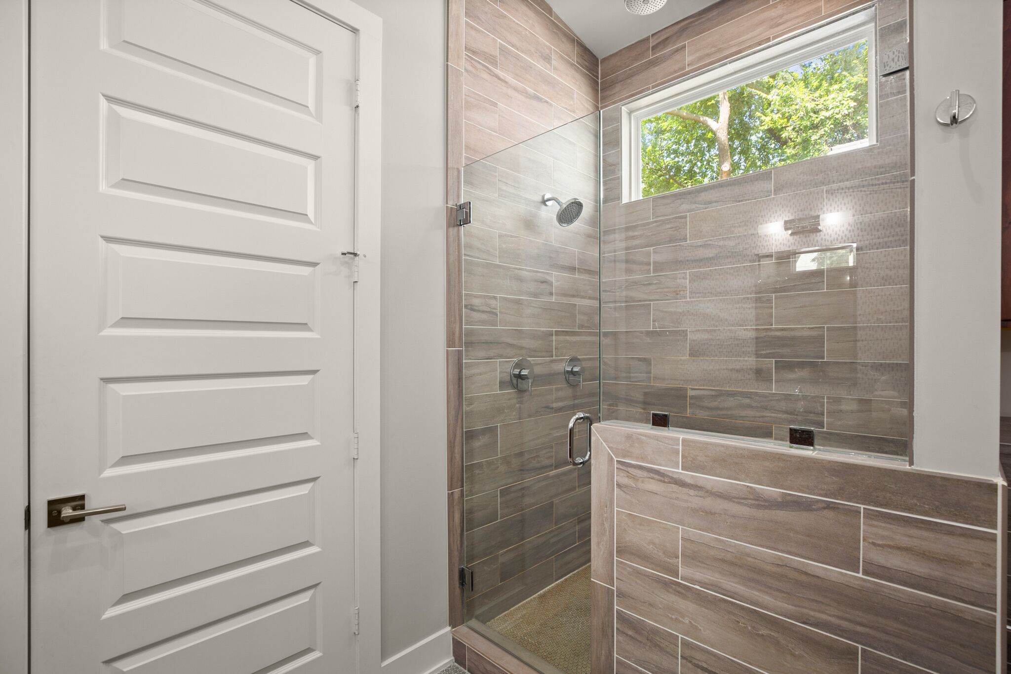 Un baño moderno con ducha a ras de suelo, una ventana con vistas a árboles y una puerta blanca.
