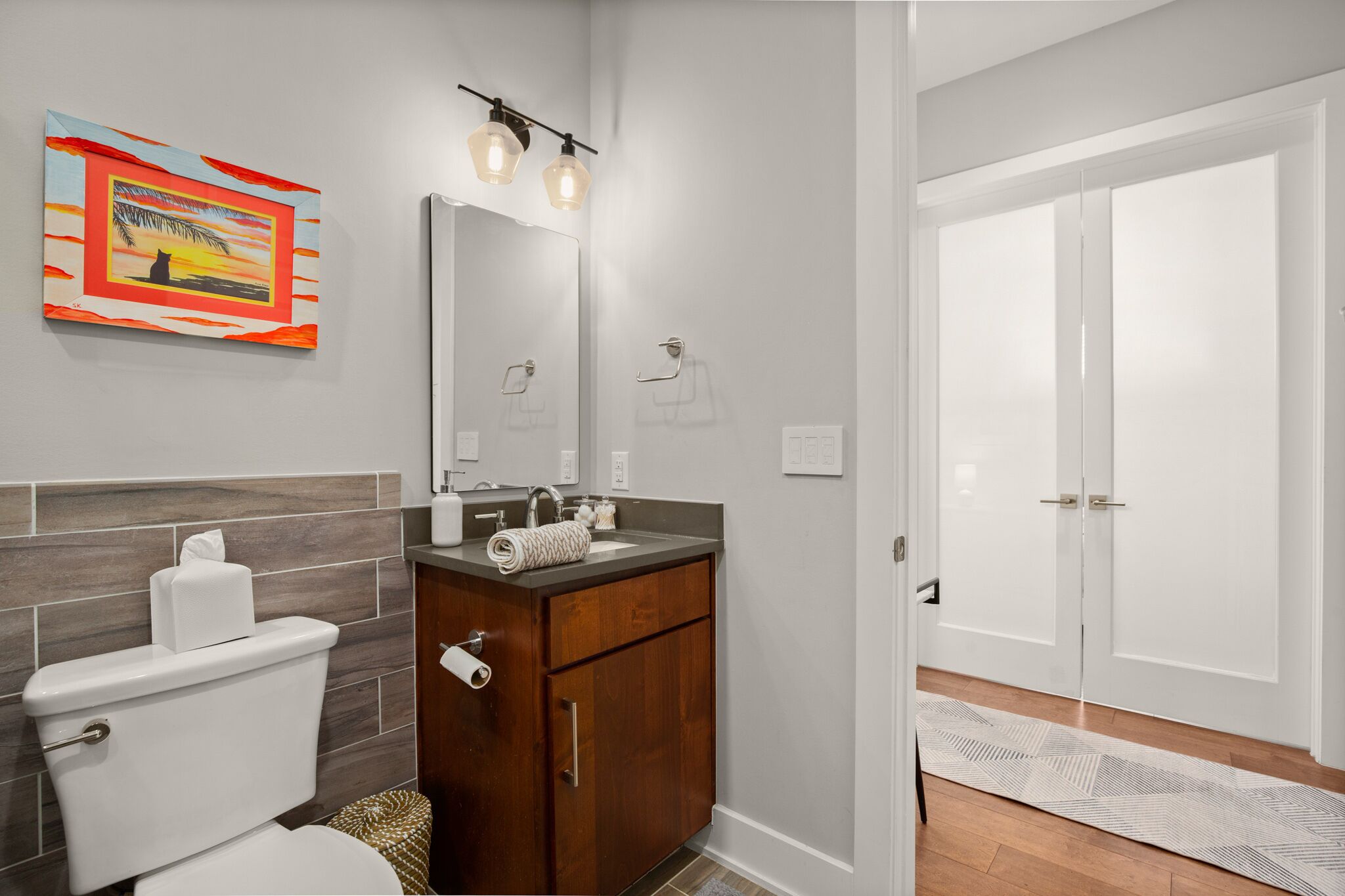 A modern bathroom with a toilet, sink, and mirror. There is a towel rack, a towel, and a toilet paper holder. The walls are tiled, and there is a door leading to another room.