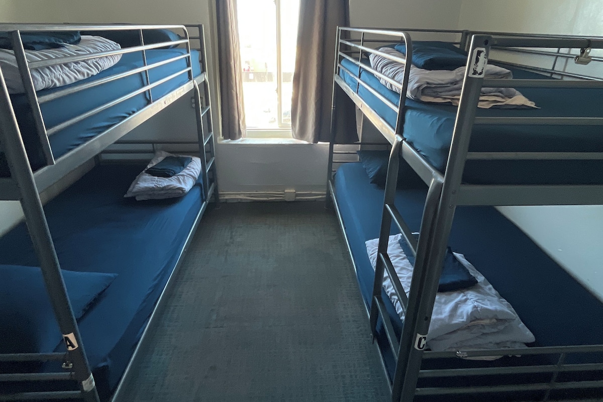 A dormitory room with bunk beds, a window with curtains, and a ceiling light.