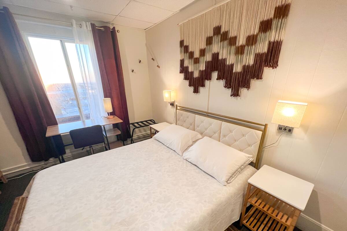 Une chambre d’hôtel avec un grand lit, un bureau avec une chaise, une petite table d’appoint en bois et une vue sur l’extérieur par une fenêtre.