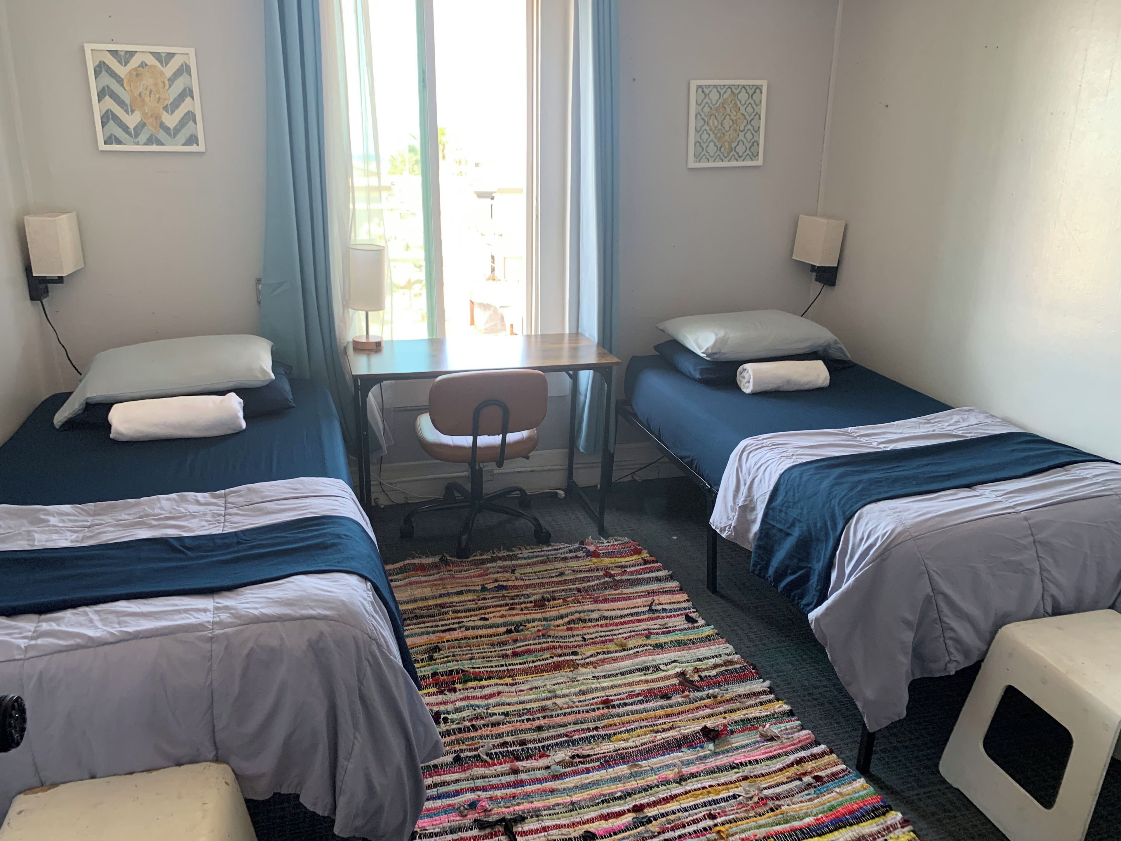 Ein Zimmer mit zwei Einzelbetten, einem Schreibtisch, einem Stuhl und einem farbenfrohen Teppich.
