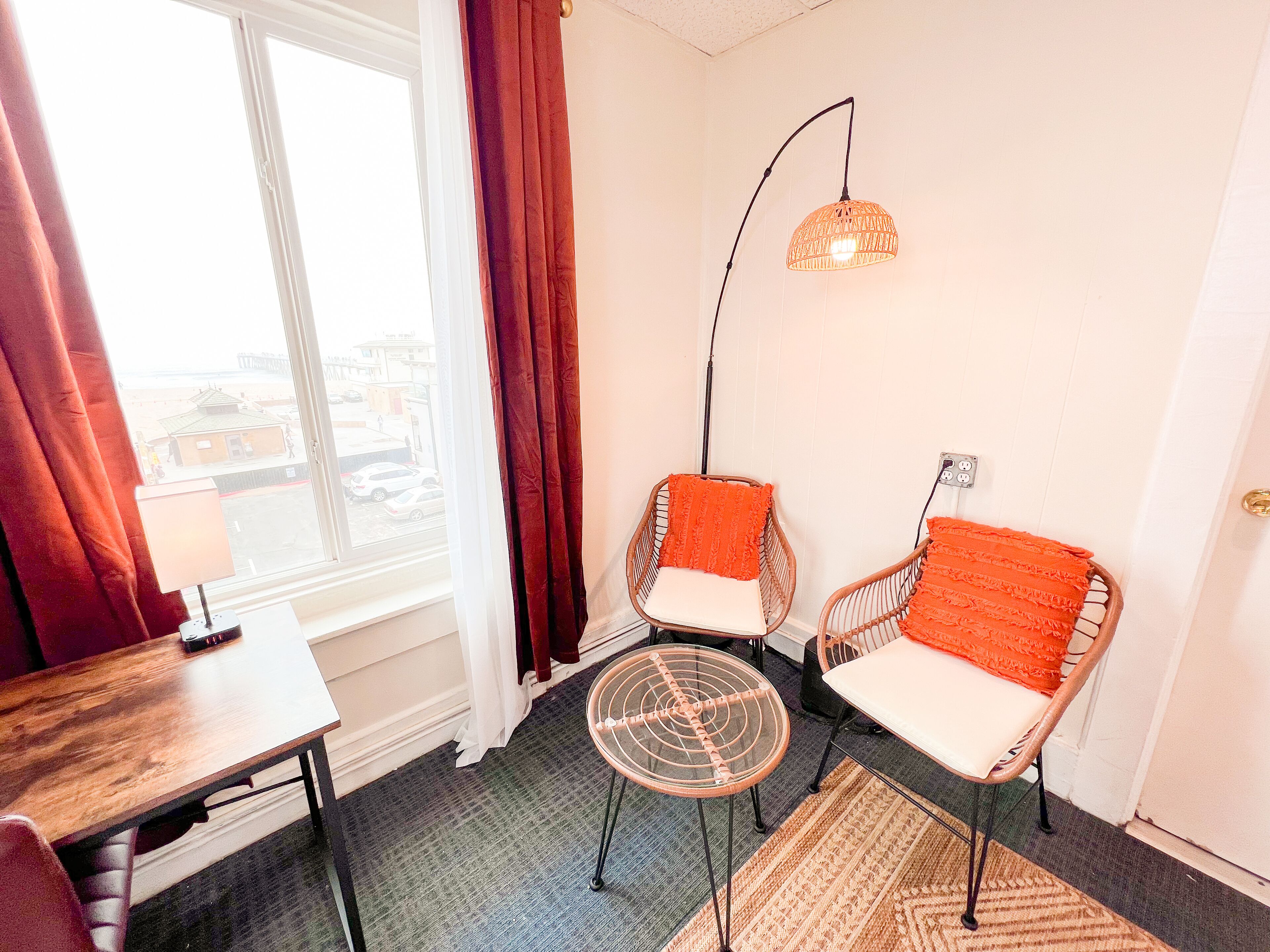 A room with two wicker chairs with orange cushions, a small round table, a floor lamp, and a view of buildings through a window with red curtains.