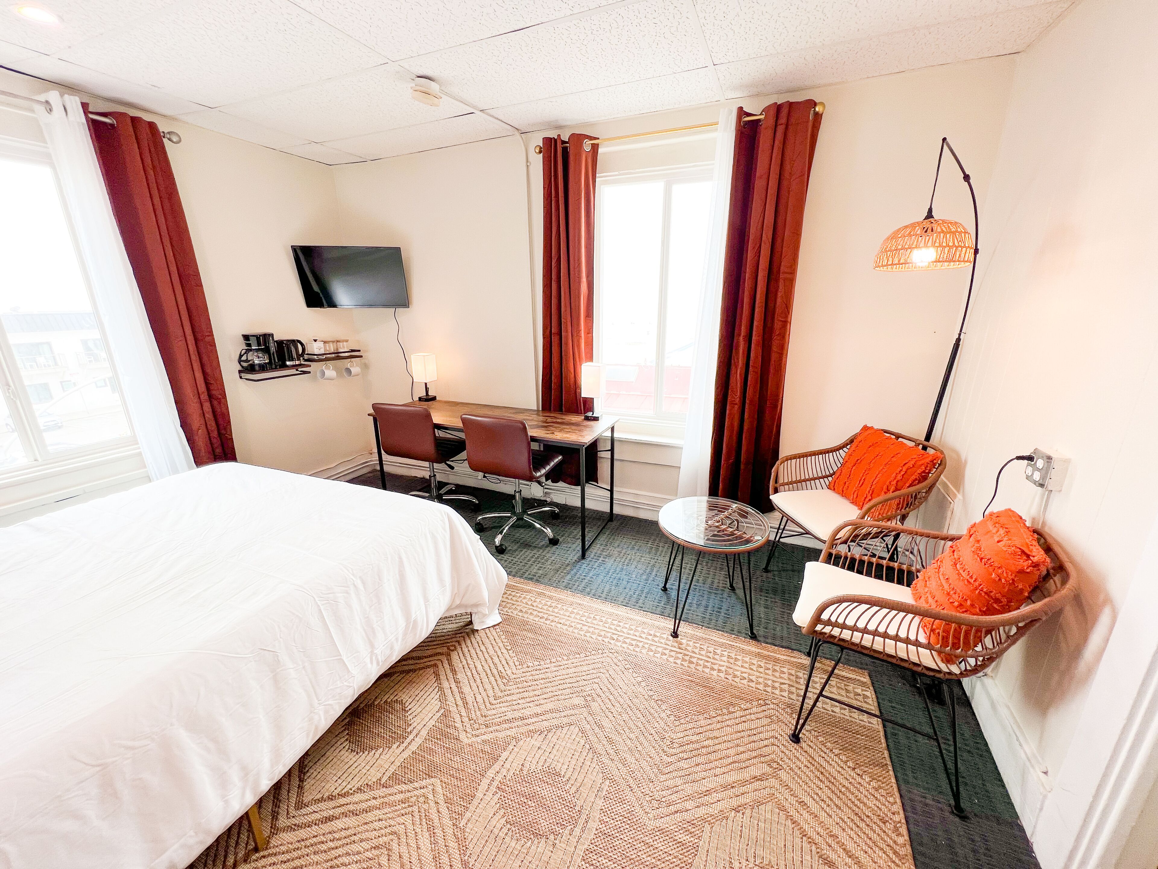 Une chambre d’hôtel comprenant un lit, un bureau avec deux chaises, une télévision, une lampe, un fauteuil en osier avec un coussin orange et un tapis à motifs.