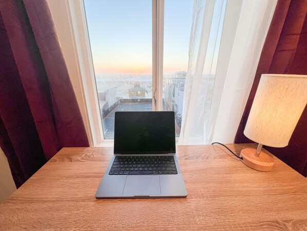 Una computadora portátil sobre una mesa de madera, junto a una ventana con cortinas, con vistas a un paisaje urbano.