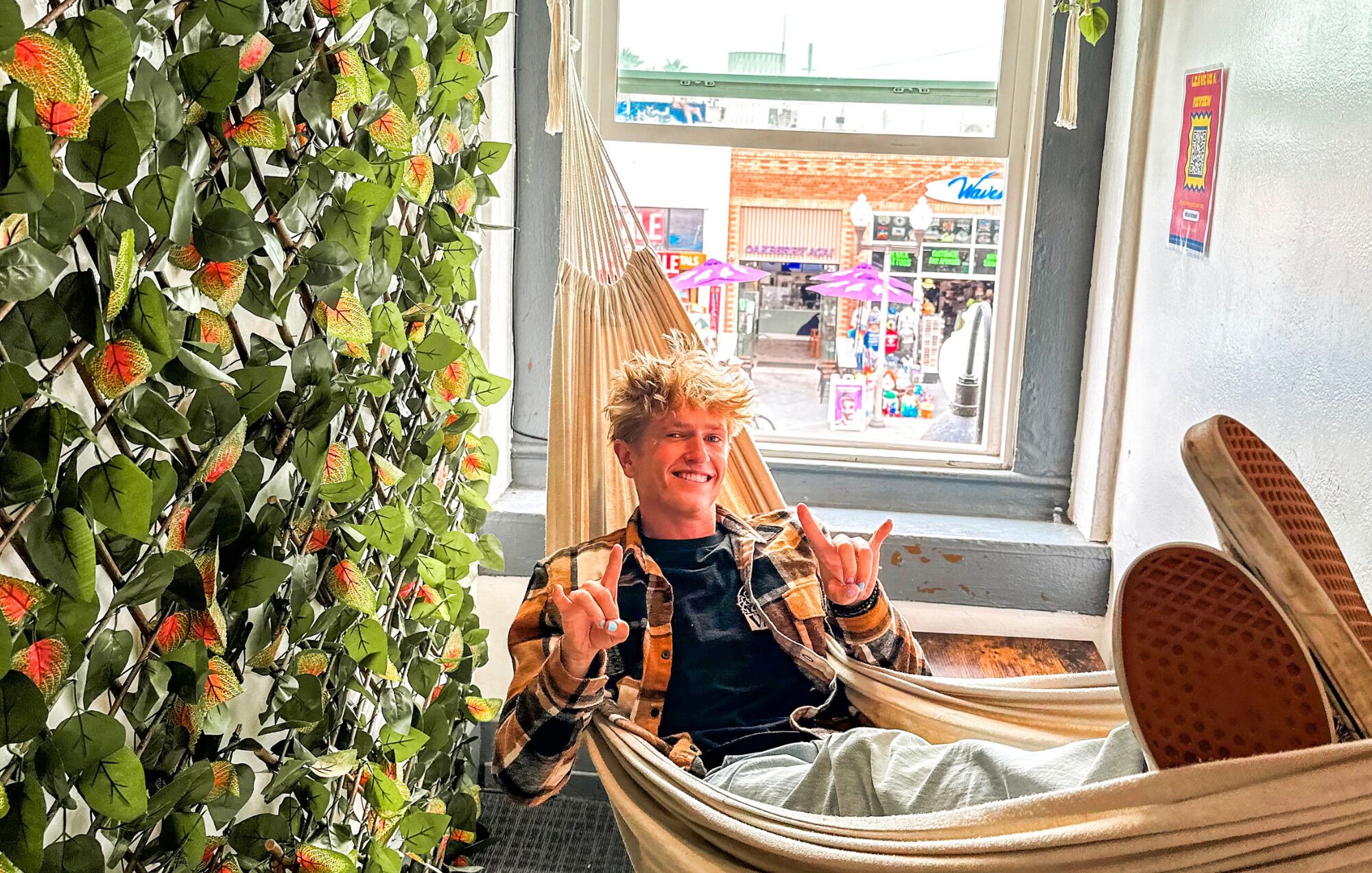 A person sitting in a hammock chair by a window with a view of a street and shops.