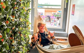 Iemand zit in een hangstoel bij een raam, met uitzicht op een straat en winkels.
