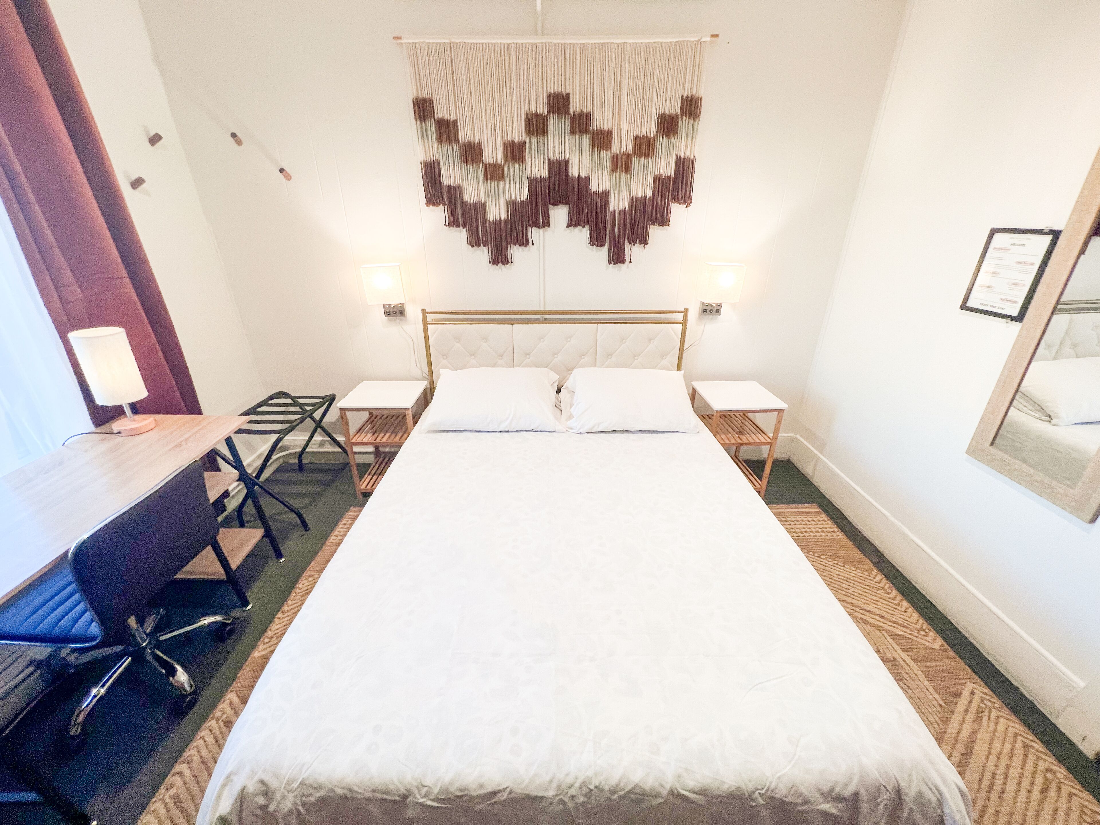 A hotel room with a large bed, two bedside tables, a desk with a chair, a wall-mounted lamp, and a decorative hanging piece.