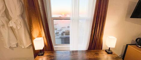 A hotel room with a wooden table, two chairs, a TV, and a view of the sunset through a window with curtains.