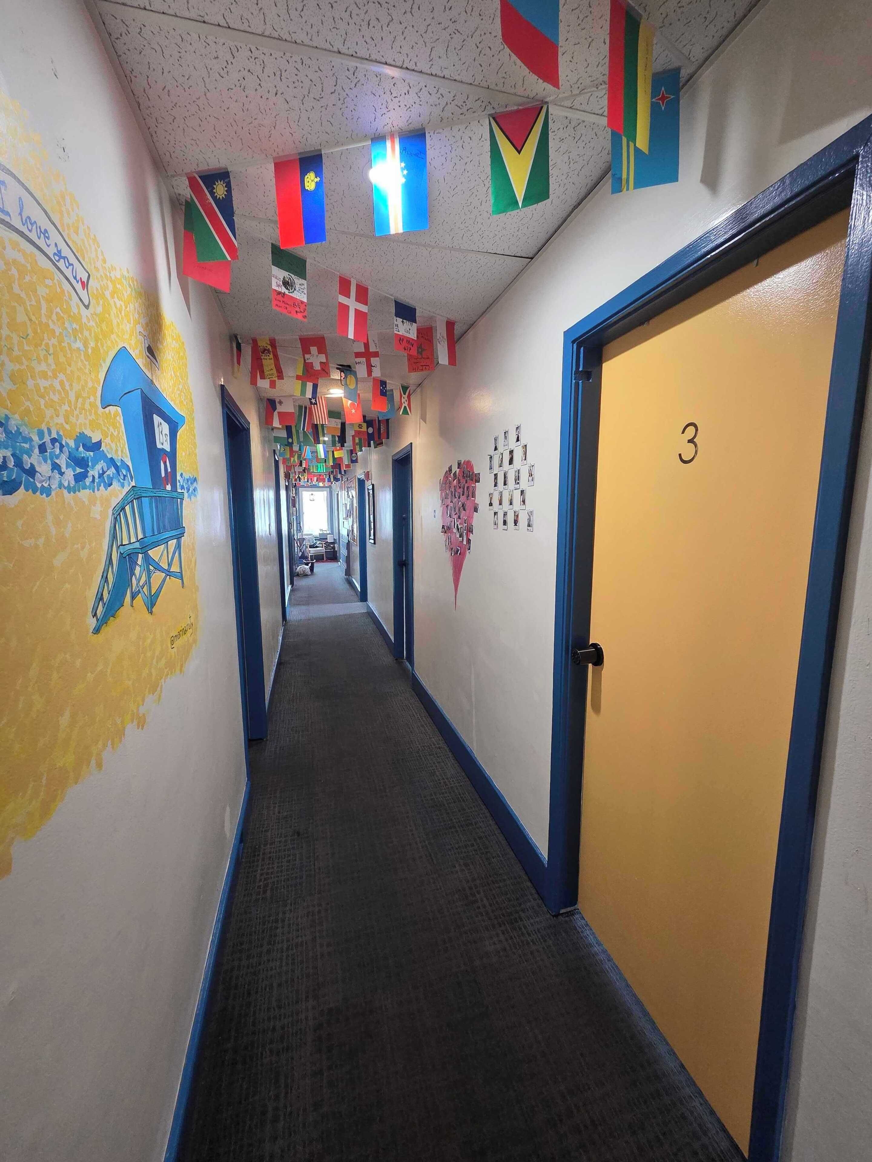 Un pasillo con piso alfombrado, una puerta azul con el número '3' y paredes decoradas con varias banderas y un collage de fotos con forma de corazón.