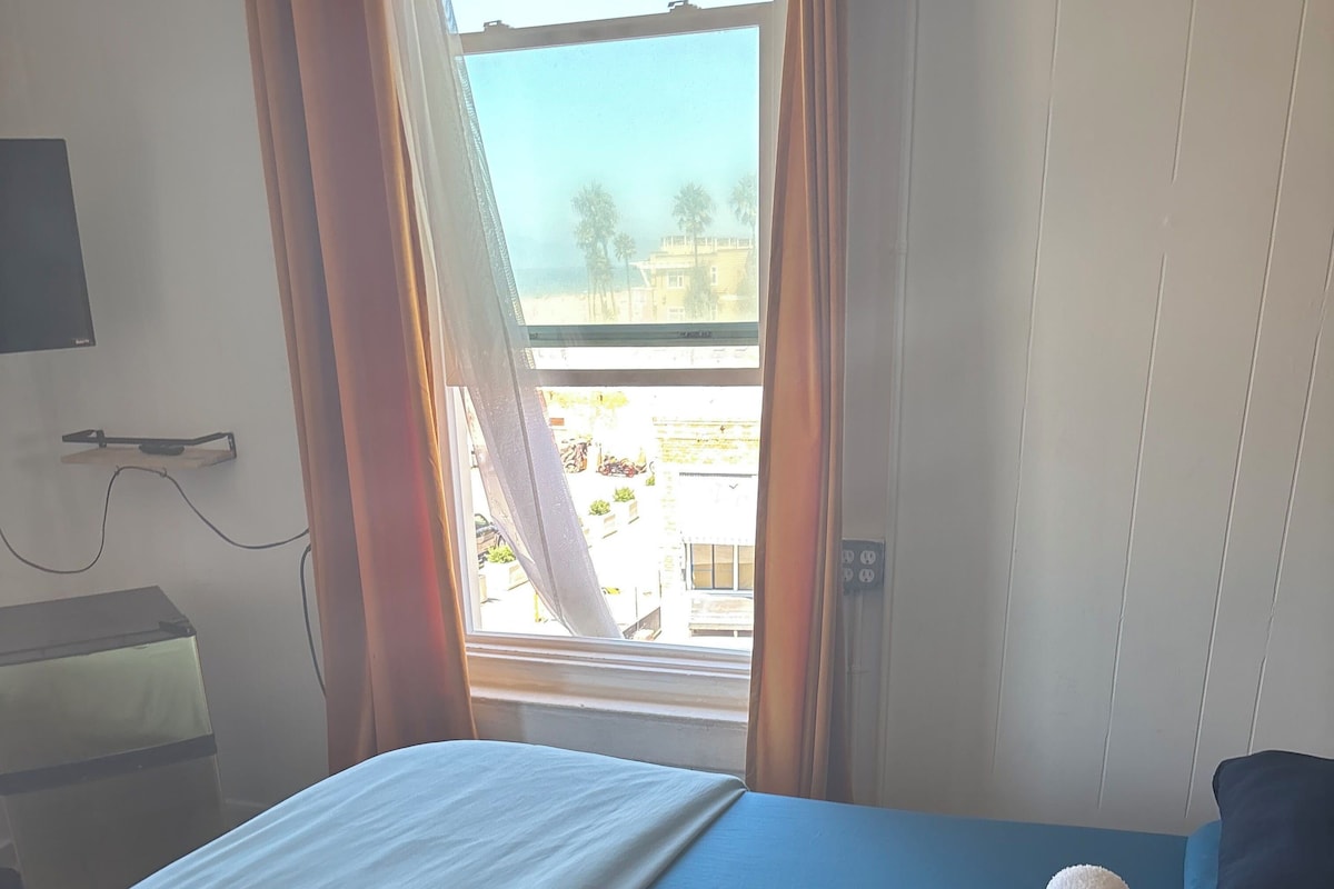 A hotel room with a bed, a window with curtains, a desk with a lamp, and a view of buildings outside.