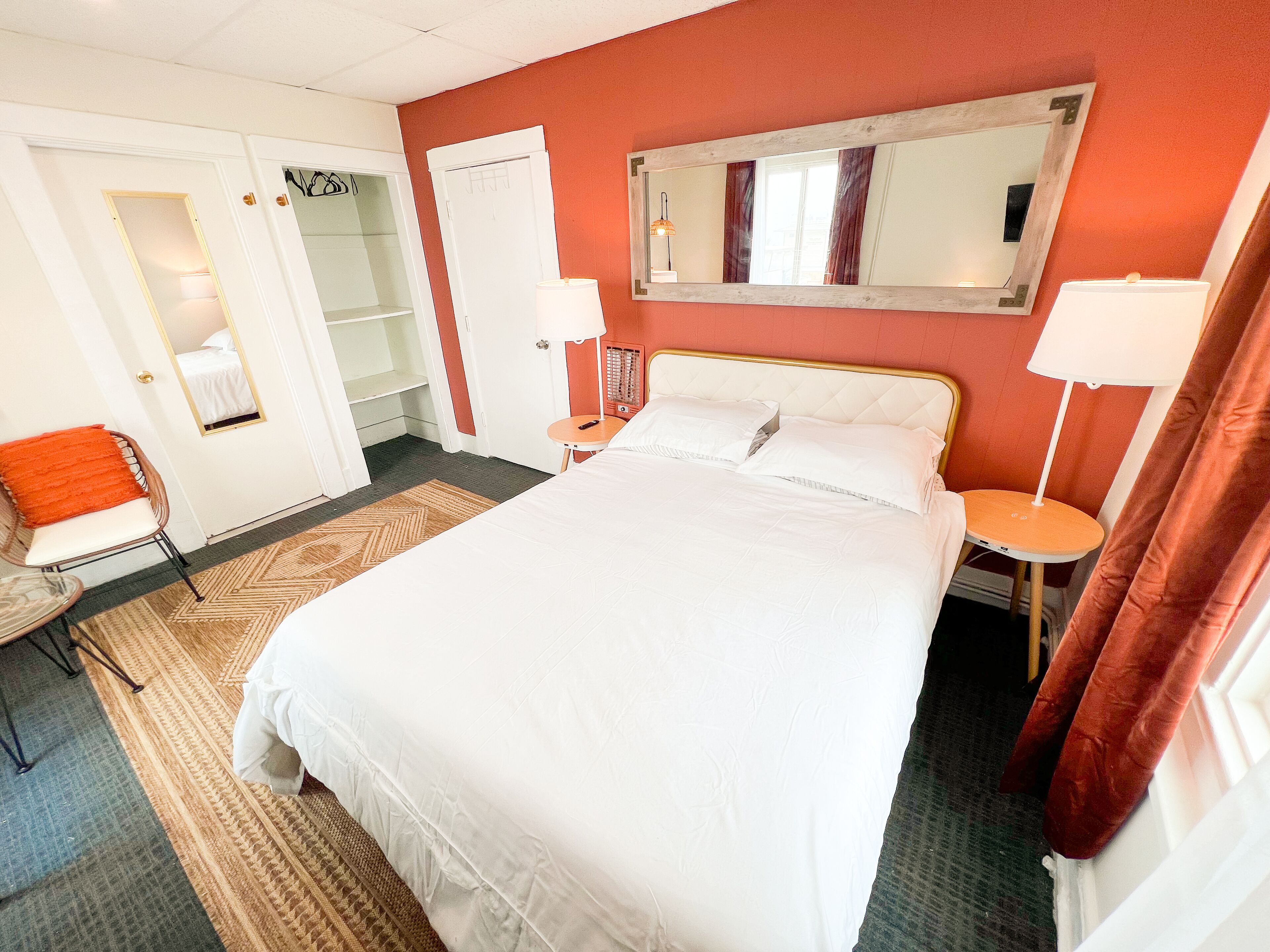 A hotel room with a large bed, a bedside table with a lamp, a chair, a wardrobe, and a mirror on the wall.