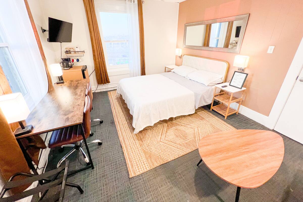 Une chambre d’hôtel avec un lit, un bureau, une chaise, une télévision, une fenêtre avec des rideaux et un miroir au mur.