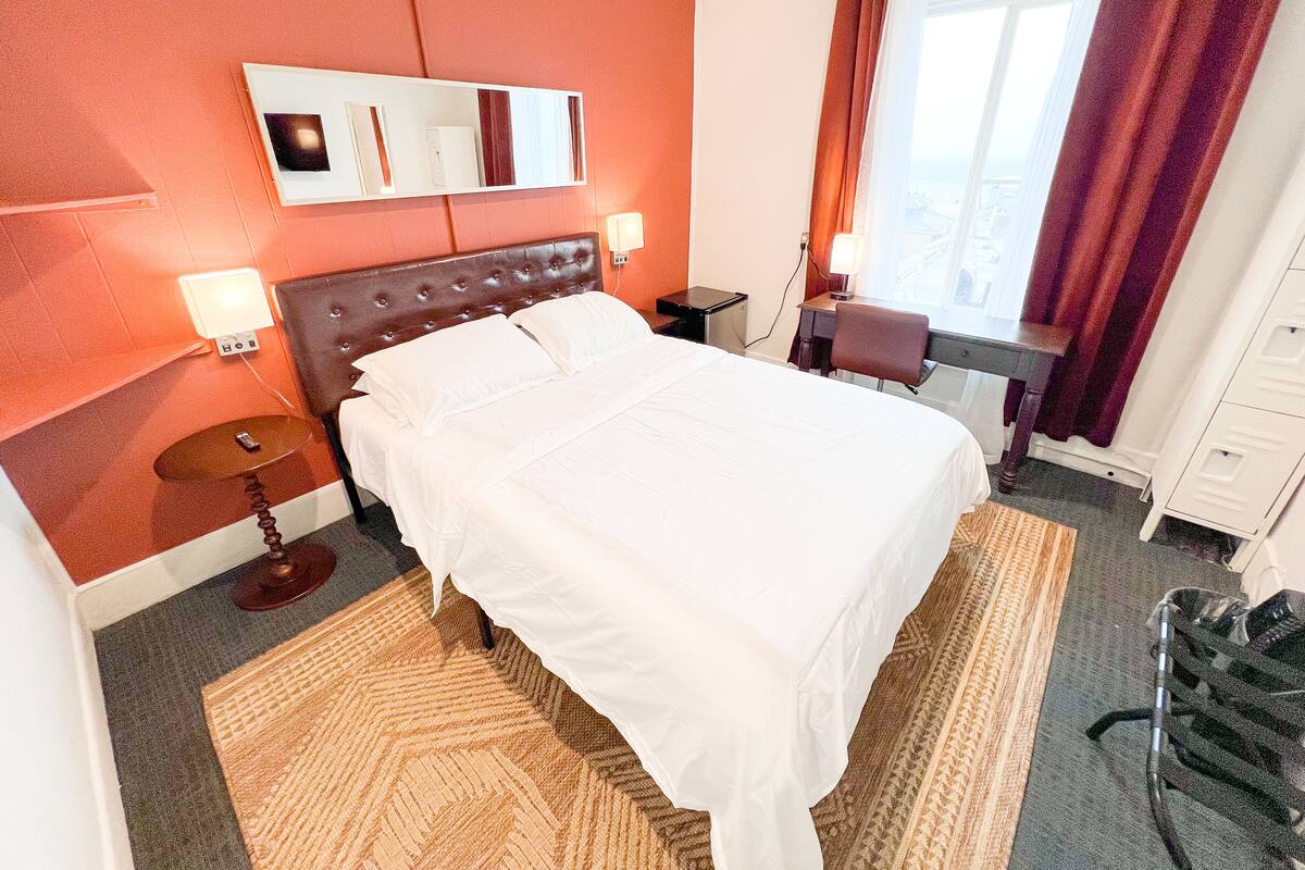 Une chambre d’hôtel avec un grand lit, un bureau, une chaise, une lampe de bureau, un téléphone et une fenêtre avec des rideaux.