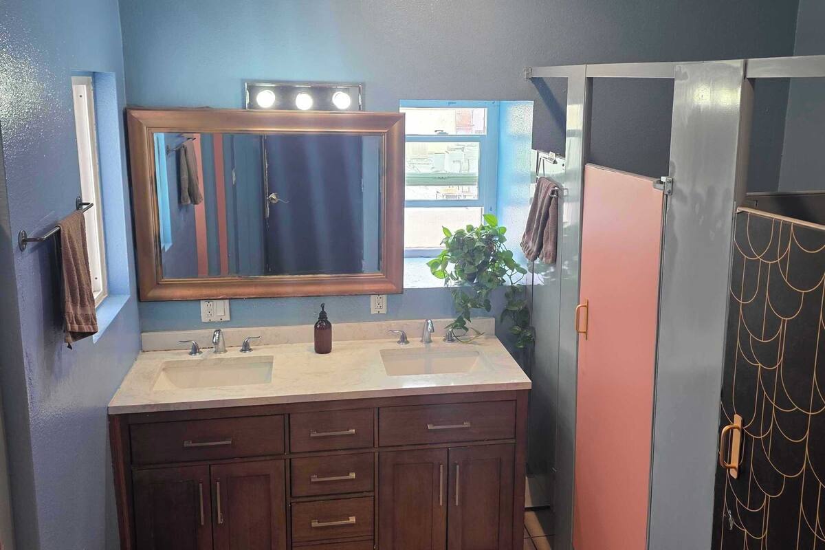 Une salle de bain équipée d’un lavabo double, d’un miroir, d’un porte-serviettes, d’une poubelle et d’une porte ornée d’un motif décoratif.