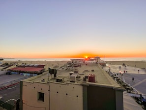 Een dakterras met uitzicht op de ondergaande zon boven de oceaan, met een parkeerplaats en terrasmeubilair.