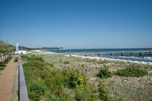 A coastal walkway with a bridge over the water, a beach, and a building with a flag.