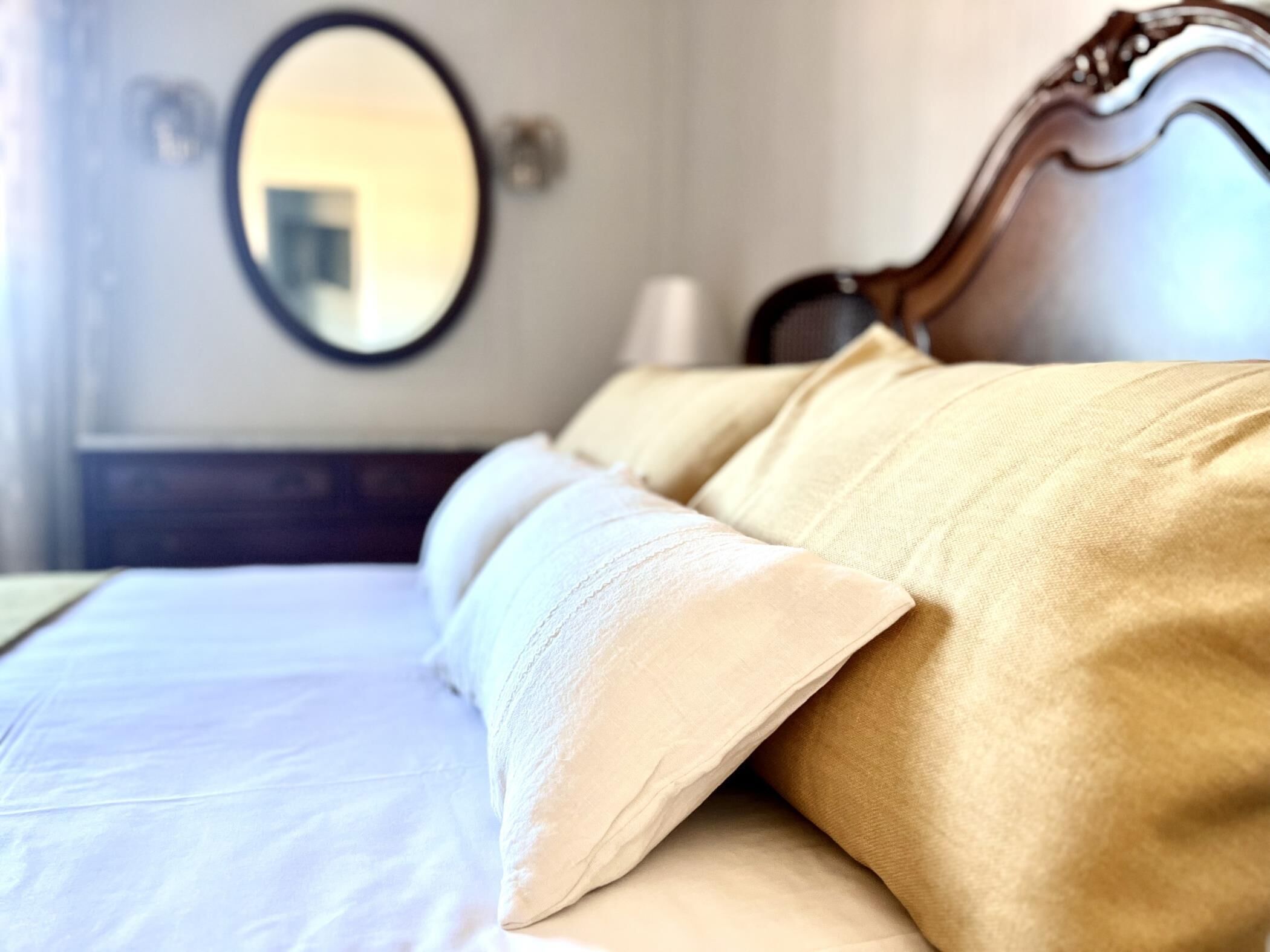 A neatly made bed with white and beige linens, a wooden headboard, and a round mirror on the wall.