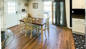 Un comedor con mesa y sillas de madera, una mesa con cubierta de vidrio y una zona de cocina con armarios blancos y encimera negra.
