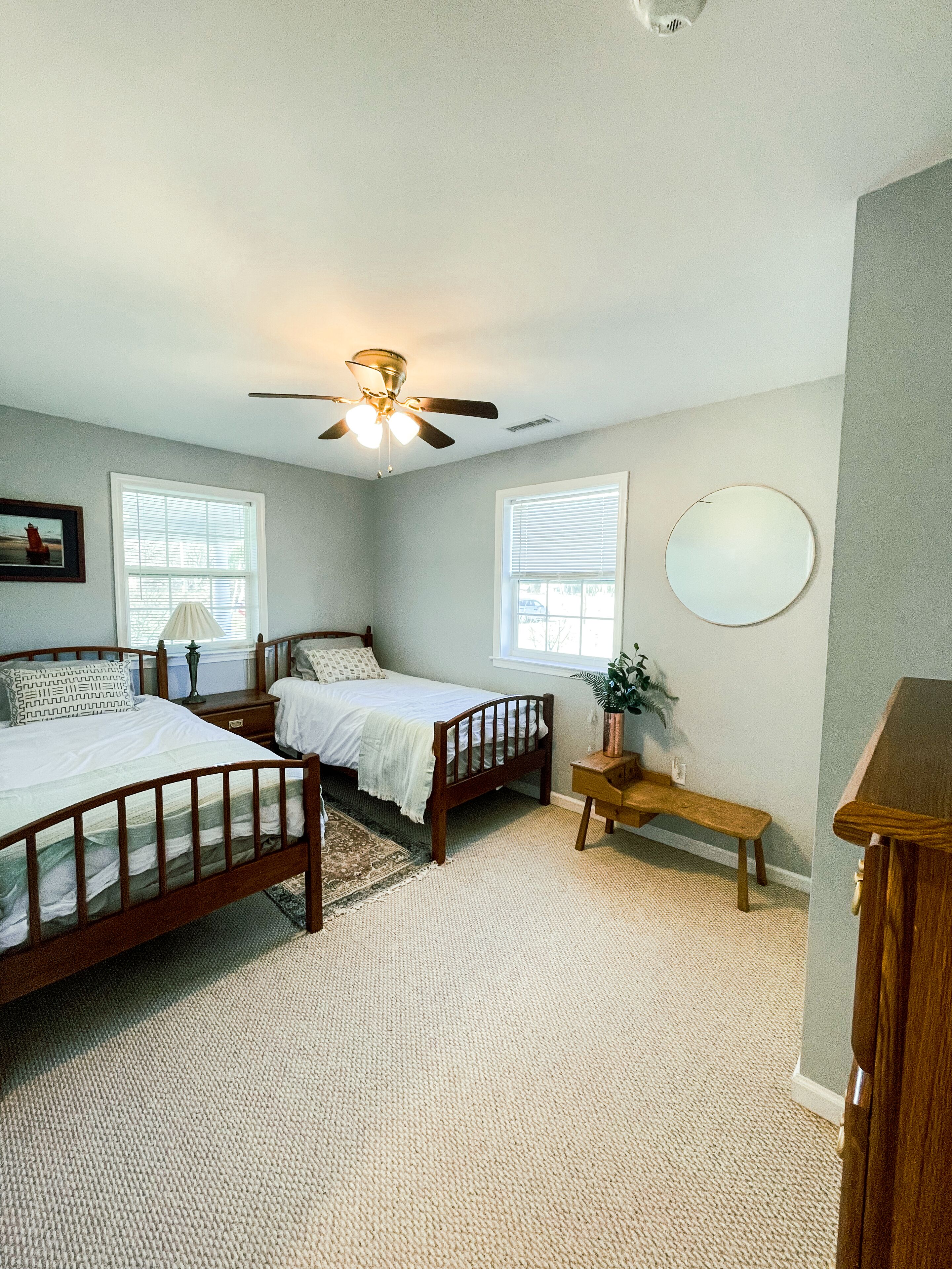 A bedroom with two beds, a ceiling fan, a bedside table with a lamp, a small bench, and a round mirror on the wall.