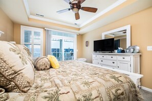Een slaapkamer met een bed, een plafondventilator, een televisie, een dressoir en een raam met jaloezieën.