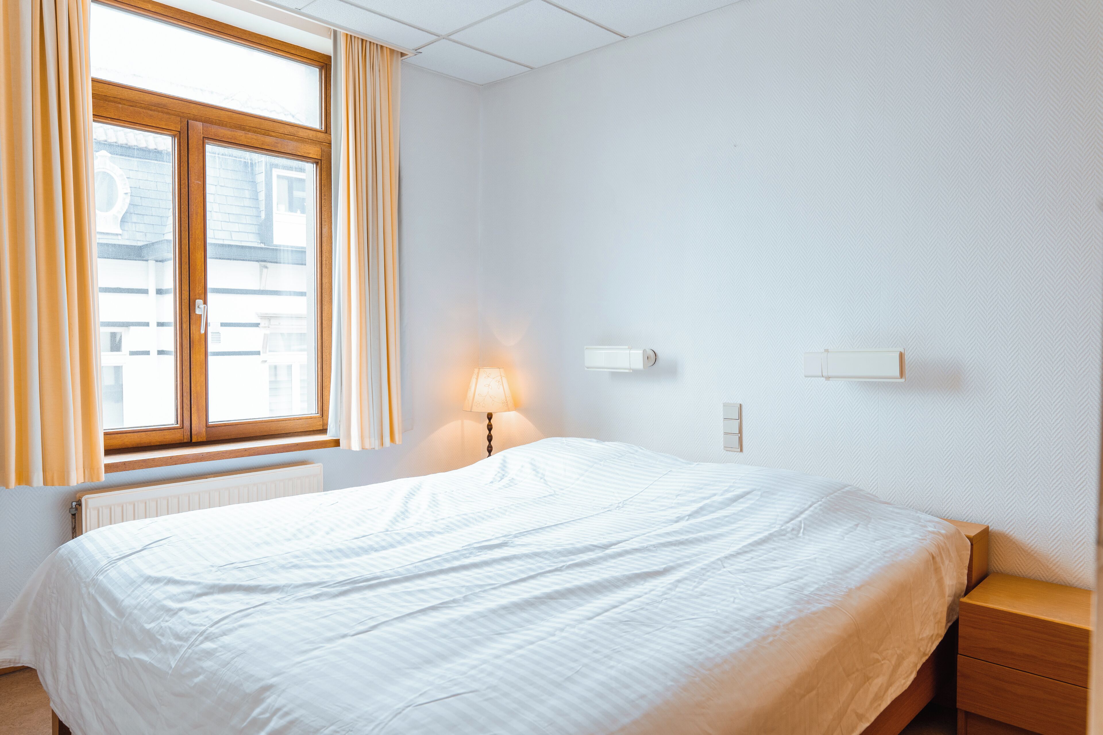 Une chambre à coucher avec un grand lit, une table de chevet, une lampe et une fenêtre avec des rideaux.