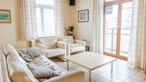 Ein Wohnzimmer mit einer Couch, zwei Sesseln und einem Couchtisch. Ein Fenster mit Vorhängen, ein gerahmtes Bild an der Wand und eine Tür, die nach draußen führt.