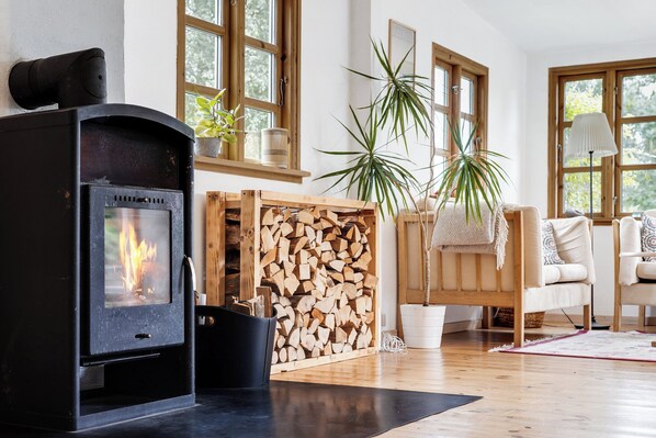 Une piĂšce chaleureuse avec un poĂȘle Ă bois, une pile de bois de chauffage, une plante en pot et une fenĂȘtre donnant sur les arbres.