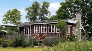 Punaisella maalilla maalattu, kaksikerroksinen talo, jossa on valkoiset listat ja savupiippu. Talon edessä on puinen terassi, jossa on kaiteet, ja taloa ympäröi puutarha, jossa kasvaa monenlaisia kasveja ja kukkia.