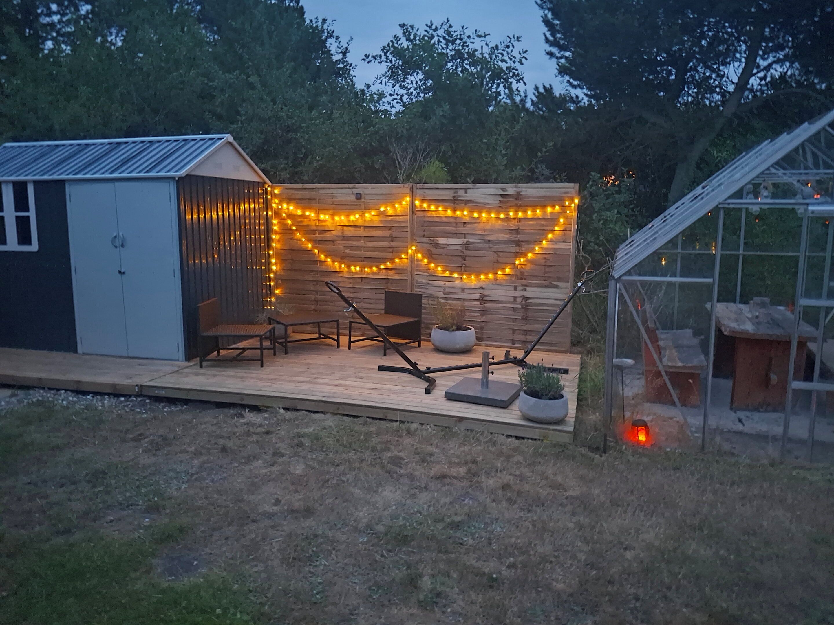 Una terraza de madera con mobiliario exterior, un cobertizo y un invernadero.
