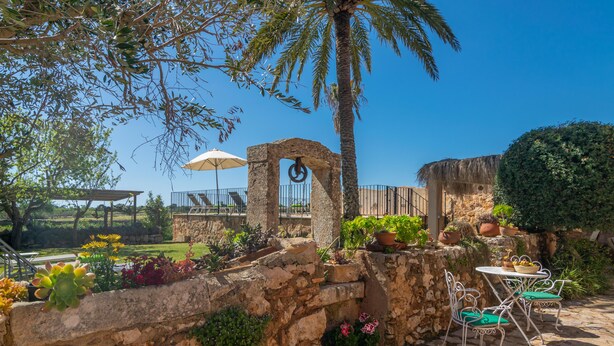 A stone wall with potted plants, a palm tree, and a metal table with chairs. - NA BURGUERA - Wonderful villa with private pool and free WiFi. (Santanyí)