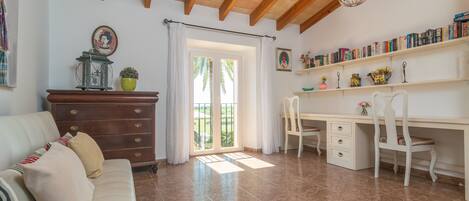 A room with a wooden ceiling, a white desk with a chair, a bookshelf, a sofa, and a window with curtains.