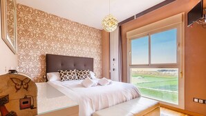 Een slaapkamer met een groot raam, een bed met witte lakens, een houten hoofdeinde en een nachtkastje met een decoratief object.